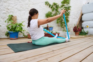 Bir kadın, dışarıda, tahta bir güvertede, bir direniş bandı kullanarak yoga yapıyor. Yeşillik tarafından kucaklanıyor. Doğal ortamlarda spor ve rahatlığı teşvik ediyor..