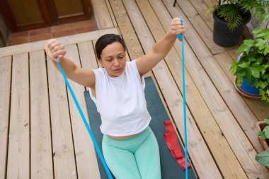 Bir kadın ahşap bir güvertede bir direniş bandıyla egzersiz yapıyor. Açık havada fitness ve sağlıklı yaşam tarzını teşvik ediyor..