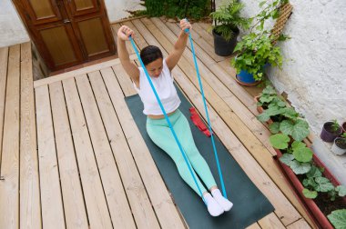 Tahta güvertede oturan bir kadın direniş bandı egzersizi yapıyor. Bitkilerle çevrili bir açık hava ortamında spor, ev çalışması ve sağlığa odaklanın..