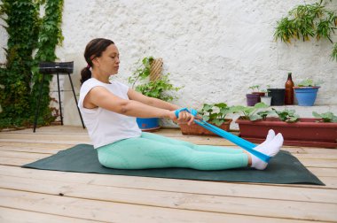 Bir kadın, çevresi yeşillik ve saksı bitkileriyle çevrili bir direniş bandıyla ahşap bir güvertede yoga yapıyor. Sağlık ve rahatlamayı teşvik ediyor..
