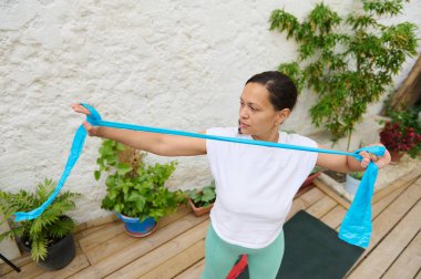 Bir kadın açık hava bahçesinde direniş bandı egzersizi yapıyor. Fitness, sağlık ve sakin çevrenin altını çiziyorum. Güç eğitimi ve refah temalı bağlamlar için ideal.