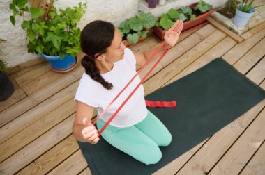 Bir kadın açık havada yoga minderi üzerinde direnç bandı egzersizleri yapıyor, yemyeşil bitkilerle dolu huzurlu bir ortamda dayanıklılık ve esnekliği arttırıyor..