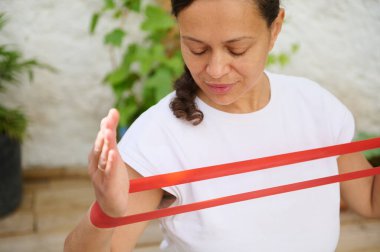 Kırmızı direnç bandını açık havada kullanan bir kadın huzurlu bir bahçe ortamında spor ve güç eğitimine odaklanmış sağlıklı yaşam tarzı ve kararlılığı somutlaştırıyor..