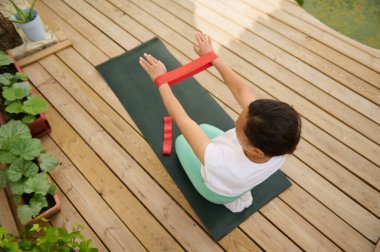 Dışarıdaki yoga minderinde egzersiz yapan bir kadının ahşap bir güvertede yemyeşil bir zeminle çevrili olduğunu görüyoruz..