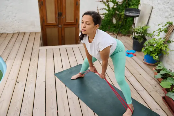 Çevresi yeşilliklerle çevrili, tahta bir güvertede direniş bandıyla yoga yapan bir kadın. Arka bahçeyi rahatlatmak, dışarıda egzersiz yaparken odak ve sükuneti arttırır..