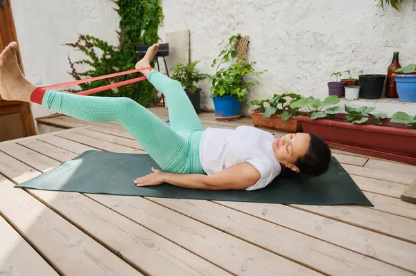 Yaşlı bir kadın ahşap bir güvertede, dayanıklılık ve sağlığını vurgulayan bir direniş bandı çalışmasına katılıyor. Etrafında saksı bitkileri ve barışçıl bir atmosfer var..