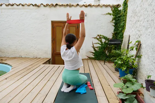 Dışarıda direnç bandıyla yoga yapan bir kadın, sakin bir bahçe ortamında güç ve esnekliğe odaklanıyor. Spor, sağlık ve açık hava egzersizleri için mükemmel..