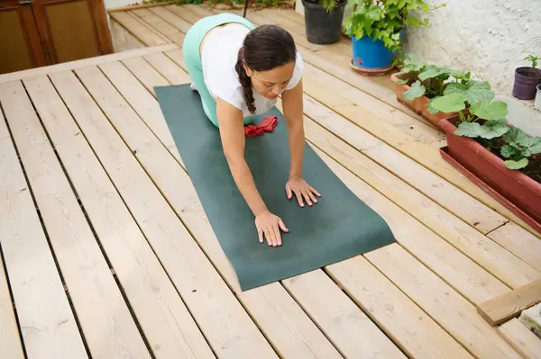 Tahta bir güvertede yoga seansına katılan bir kadın, yeşil bir paspas kullanıyor ve çeşitli bitkilerle çevrili, sakin ve konsantre bir şekilde.