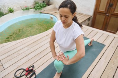 Bir kadın, dışarıda, tahta bir güvertede, rahat spor kıyafetleri giymiş, rahat bir havuzun yanında yoga hamlesi yapıyor. Huzurlu bir ortamda rahatlama ve zindelik deneyimi.