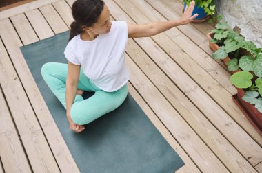 Beyaz gömlekli ve deniz mavisi taytlı bir kadın ahşap bir güvertede yoga yapıyor. Huzurlu bir ortamda rahatlama, farkındalık ve açık hava sağlığını teşvik ediyor..