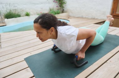Bir kadın dışarıda yoga minderinde şınav çekiyor, dayanıklılık ve zindelik üzerine odaklanıyor. Açık hava idman ortamında destek için şınav çubukları kullanıyor..