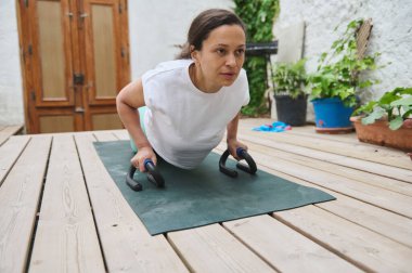 Odaklanmış bir kadın, tahta bir güvertede parmaklıklarla şınav çekiyor, spor yapmayı kucaklıyor ve dışarıda sağlıklı bir yaşam sürüyor..
