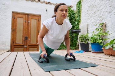 Atletik giyinmiş, yoga minderinin üzerinde şınav çeken açık havada şınav çeken, etrafı saksı bitkileri ve ahşap güverte ile çevrili odaklanmış bir kadın..