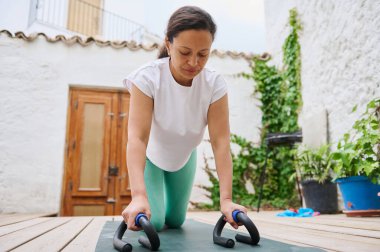 Odaklanmış bir kadın, açık avluda bir paspasın üzerinde şınav çekiyor, fitness ve kuvveti destekliyor..