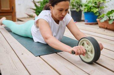 Bir kadın, dışarıda yoga minderinin üzerinde bir kas silindiri ile vücut egzersizi yapıyor. Huzurlu bir bahçe ortamında gücünü ve sağlığını arttırıyor..