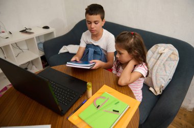 İki çocuk evde derslerine odaklanıyor, masada kitap ve dizüstü bilgisayarla uğraşıyorlar..