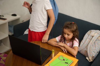 Two children focused on an online lesson at home. A girl sits attentively at a table, while a boy stands nearby. Notebooks and school supplies are visible, emphasizing education. clipart