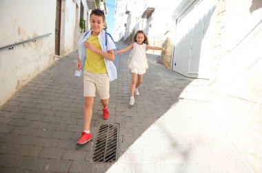 Sırt çantalı iki neşeli çocuk parlak, güneşli bir sokakta koşuşturuyorlar, kaygısız bir oyun anının tadını çıkarıyorlar..