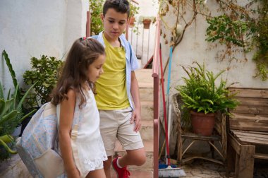 Sırt çantalı iki çocuk, güneşli bir ortamda, arkadaşlığı ve macerayı simgeleyen bitkilerle çevrili olarak merdivenlerden aşağı iniyor..