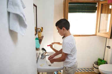 Genç bir çocuk banyo lavabosunda dişlerini fırçalıyor. Parlak, aydınlık bir alanda kişisel hijyene vurgu yapıyor..