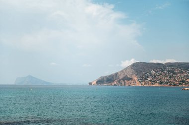 Huzurlu bir denizi, engebeli dağ zeminini ve açık mavi gökyüzünü barındıran resmedilmeye değer kıyı manzarası. Seyahat ve doğa temaları için ideal.