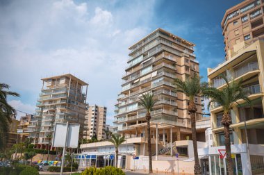 Parlak mavi bir gökyüzünün altında, palmiye ağaçlarıyla çevrili, balkonları olan modern apartman binalarının yer aldığı şehir manzarası. Gayrimenkul veya şehir manzarası temaları için ideal.