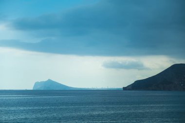 Huzurlu deniz manzarası, bulutlu gökyüzü, uzak dağlık kıyı şeridi ve ufukta şehir silüeti..