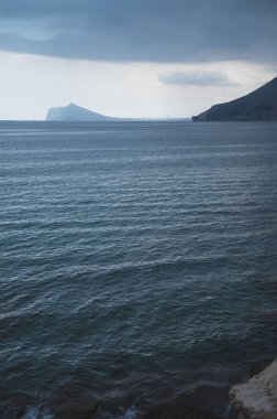 Bulutlu gökyüzünün altında sakin mavi suları resmeden sakin bir deniz manzarası ufukta uzak dağlar, sakin ve karamsar bir atmosfer yaratıyor..