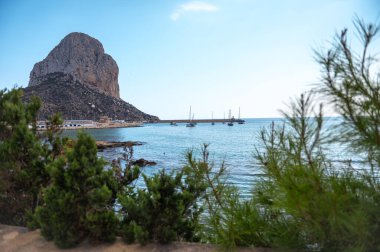 İspanya 'nın Calpe kentindeki Penon de Ifach manzaralı sakin sular ve yelkenliler. Bereketli yeşillik, huzurlu Akdeniz kıyı şeridini çevreleyerek mükemmel bir yaz kaçışı sağlıyor..