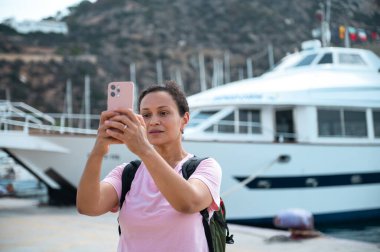 Pembe tişörtlü bir kadın, resim gibi bir limanda bir yatın yanında selfie çekiyor. Seyahat macerasından zevk alıyor..