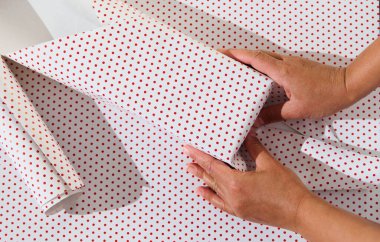 Eller kırmızı benekli kağıtla bir hediyeyi dikkatlice paketliyor, hediye hazırlama sanatını ve şenlikli ruhu vurguluyor..