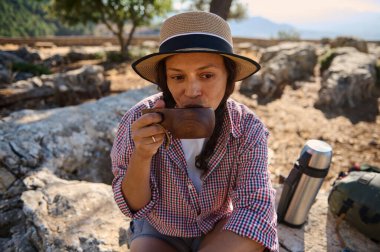 Bir kadın, doğa güzelliğiyle çevrili hasır bir şapka ve ekose gömlek giyerek, huzurlu bir doğa yürüyüşü sırasında sıcak bir içki içmekten hoşlanır..