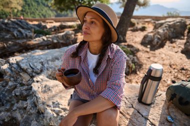 Kayalıklarda oturan şapkalı bir kadın, dışarıda bir fincan kahvenin tadını çıkarıyor. Doğal ortamda sakin ve huzurlu bir an..