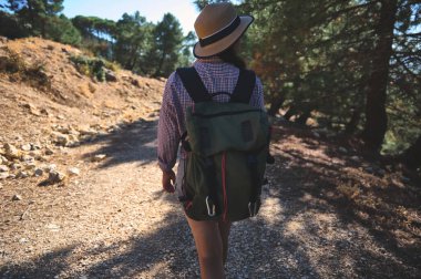 Ağaçlarla çevrili güneşli bir orman patikasında şapka ve sırt çantası taşıyan bir kadın. Macera ve sükuneti simgeliyor..