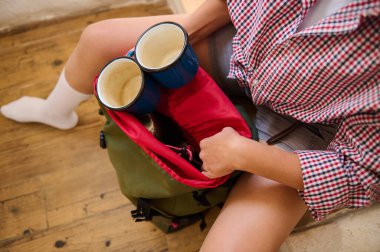 Bardakları sırt çantasına doldurup açık hava macerasına hazır birinin üst görüntüsü. Gündelik kıyafetler ve parlak renkler, rahat ve açık hava havasını vurgular..