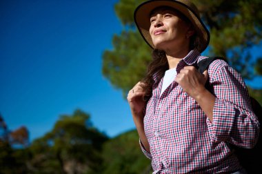 Kendine güvenen bir kadın güneşli bir açık havada yürüyüş yapmaktan hoşlanır. Bir şapka ve sırt çantası takıyor, mutlu ve maceraperest görünüyor. Mavi gökyüzü huzur ortamını daha da güzelleştiriyor.