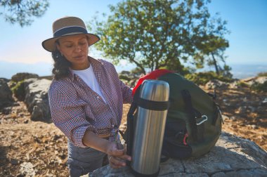 Şapka ve kareli gömlek giymiş bir kadın yürüyüş için hazırlanıyor, termoslu bir sırt çantasının yanında duruyor. Manzaralı dağ manzarası ve güneşli hava açık hava macerasını güçlendirir..