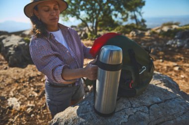 Bir kadın dağ yürüyüşü sırasında, sırt çantasının yanında bir termos tutarak mola verir. Barışçıl dış mekan doğaya ve maceraya olan sevgiyi vurguluyor..