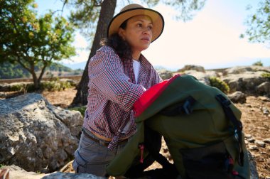 Şapkalı ve kareli gömlekli bir kadın etrafı kayalar ve ağaçlarla çevrili sırt çantasıyla dışarıyı keşfediyor. Resim maceranın özünü ve doğayla bağlantısını yakalar..