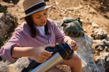 Yürüyüş sırasında şapka takan bir kadın termostan sıcak bir içecek dolduruyor. Ortam doğal ve kayalık bir manzarayla dolu. Rahatlatıcı ve ferahlatıcı bir an..