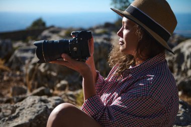 Hasır şapkalı ve ekose gömlekli bir kadın elinde kamerayla dışarıda oturuyor. O, kayalık manzarayı fotoğraflarken doğanın tadını çıkarıyor. Bir macera ve keşif hissi..