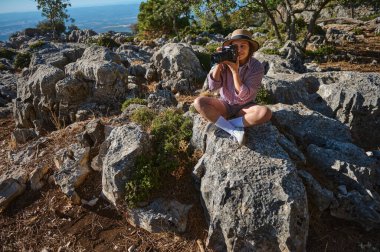 A woman sits on rocky terrain, capturing moments with a camera, embracing nature's beauty under sunlight. clipart