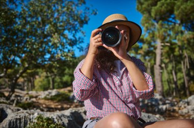 Şapka takan bir kadın kamerayı tutuyor, yemyeşil bir ormanda anları yakalıyor. Aydınlık gün, doğadaki fotoğrafçılık tutkusunu vurguluyor. Keşif ve yaratıcılığın bir resmi.