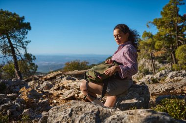 Bir kadın yürüyüş sırasında bir kayanın üzerinde oturuyor, sakin dağ manzarasının tadını çıkarıyor. Sırt çantası açık, ve bu canlı doğal ortam huzur ve macera duygusunu arttırıyor..