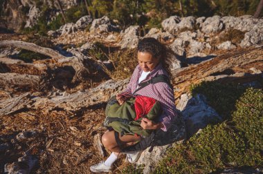 Bir kadın, ağaçlarla çevrili engebeli, kayalık bir arazide yürüyüş yaparken bir kayanın üzerinde oturup sırt çantasına bakarken huzurlu bir anın tadını çıkarır..