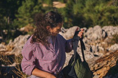 Ekose gömlek giyen bir kadın, doğa ve güneş ışığıyla çevrili kayalık bir arazide sırt çantasıyla ayakta duruyor..