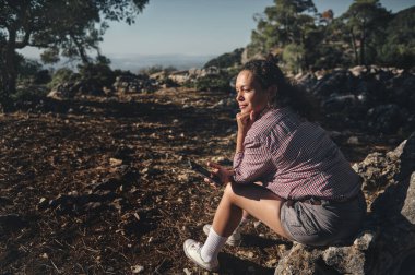 Dışarıdaki manzaralı bir kayanın üzerinde oturan bir kadın, huzurlu doğanın tadını çıkarıyor. Akıllı bir telefon tutuyor ve açık mavi gökyüzünün altında derin bir düşünce ya da tefekküre dalmış gibi görünüyor..