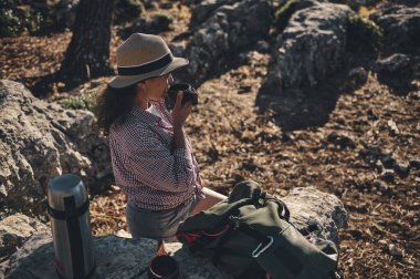 Doğayla çevrili bir kayanın üzerinde otururken şapkalı genç bir kadın kahvesini yudumluyor. Yakınlarda bir sırt çantası ve termosla öylesine giyinmiş, huzurlu bir yürüyüş anının tadını çıkarıyor..