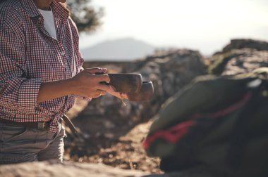 Ekose gömlek giyen bir insan, açık havada kahvenin tadını çıkaran ahşap bir fincan tutar. Atmosfer macera, rahatlama ve doğa değerini taşır..