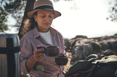 Şapkalı bir kadın, doğada kahve molası verip, dışarıda kahve içmekten hoşlanır. Etrafı ağaçlar ve kayalarla çevrili, rahatlama ve sükunet yayıyor. Seyahat ve eğlence konuları için ideal..